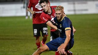 Zwei Fußballspieler führen einen körperlichen Kampf auf dem Feld, einer in Rot und der andere in Blau. Der Spieler in Rot hat seinen Arm um den Spieler in Blau gelegt, der versucht, sich zu befreien. Im Hintergrund steht ein weiterer Spieler mit der Hand auf der Hüfte.
