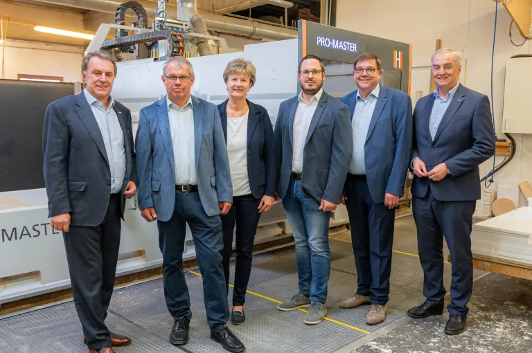 Fünf Erwachsene stehen in einer Reihe vor einer Pro-Master-Maschine und lächeln für die Kamera. Sie sind in formeller Kleidung gekleidet. Hinter ihnen ist eine große Maschine zu sehen, mit einem orangefarbenen und schwarzen Schild, auf dem Pro-Master steht.