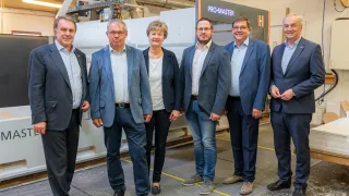 Fünf Erwachsene stehen in einer Reihe vor einer Pro-Master-Maschine und lächeln für die Kamera. Sie sind in formeller Kleidung gekleidet. Hinter ihnen ist eine große Maschine zu sehen, mit einem orangefarbenen und schwarzen Schild, auf dem Pro-Master steht.