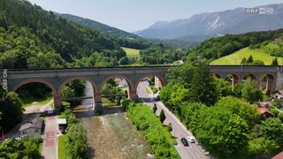 Eine Luftaufnahme eines Flusses mit einer Brücke und einer Straße darunter. Bäume und Berge sind im Hintergrund.