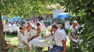 Eine Gruppe von Menschen versammelt sich unter einem Regenschirm bei einer Veranstaltung im Freien, sitzt um Tische mit Essen und Getränken, gekleidet in Weiß und Freizeitkleidung, umgeben von Grün und Blumen.