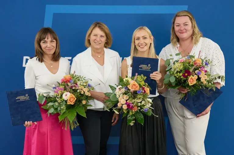 Fünf Frauen in weißen Kleidern posieren mit Blumensträußen für ein Foto. Sie stehen vor einem blauen Hintergrund. Die Frau in der Mitte hält eine blaue Mappe.