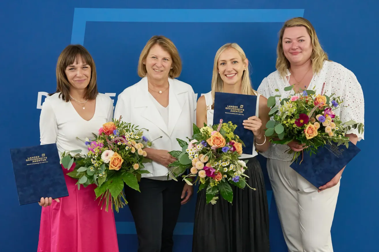 Fünf Frauen in weißen Kleidern posieren mit Blumensträußen für ein Foto. Sie stehen vor einem blauen Hintergrund. Die Frau in der Mitte hält eine blaue Mappe.