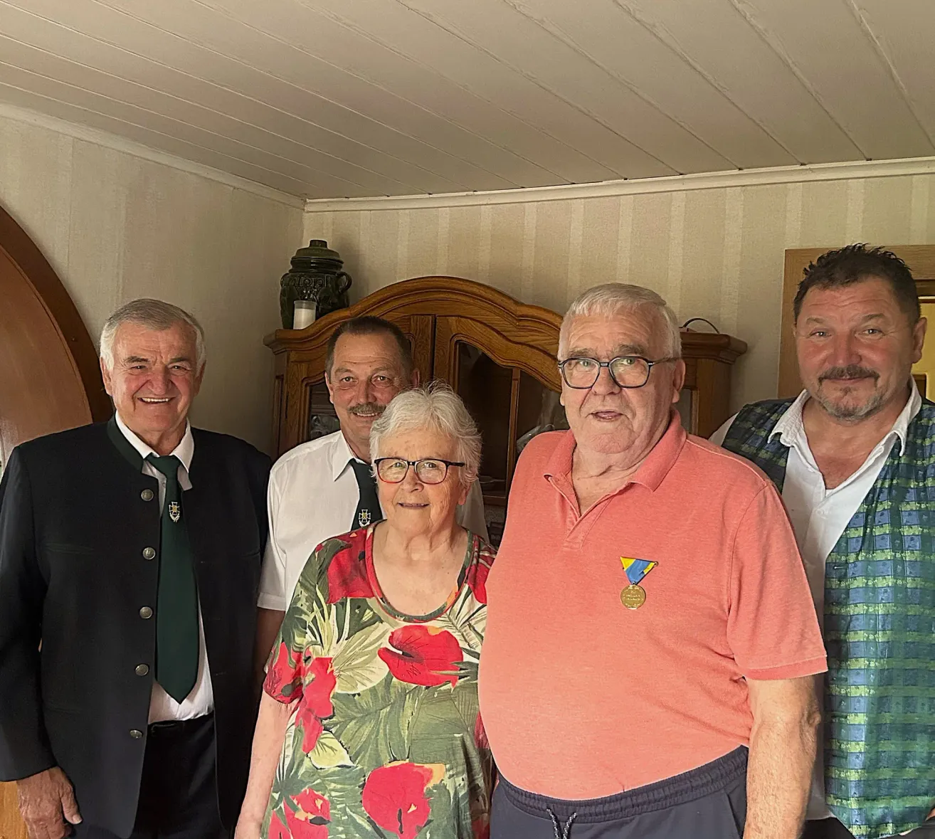 Fünf Menschen stehen lächelnd für ein Foto in einem Raum. Eine Frau mit Brille und einem Blumentop steht in der Mitte. Männer auf beiden Seiten tragen Krawatten und Westen.