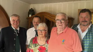 Fünf Menschen stehen lächelnd für ein Foto in einem Raum. Eine Frau mit Brille und einem Blumentop steht in der Mitte. Männer auf beiden Seiten tragen Krawatten und Westen.