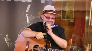 Ein Mann mit Hut und Brille spielt Gitarre und Mundharmonika in ein Mikrofon vor einem Banner.