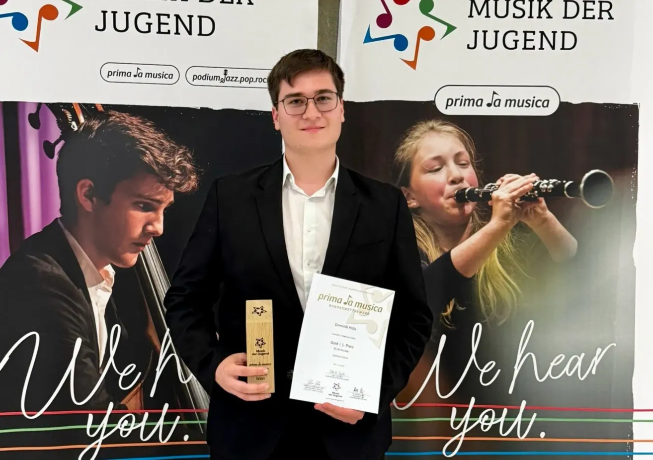 Ein junger Mann in Anzug hält einen Musikpreis und ein Zertifikat. Hinter ihm spielt eine Frau Klarinette. Der Hintergrund zeigt Musikinstrumente und den Text 'prima la musica'.