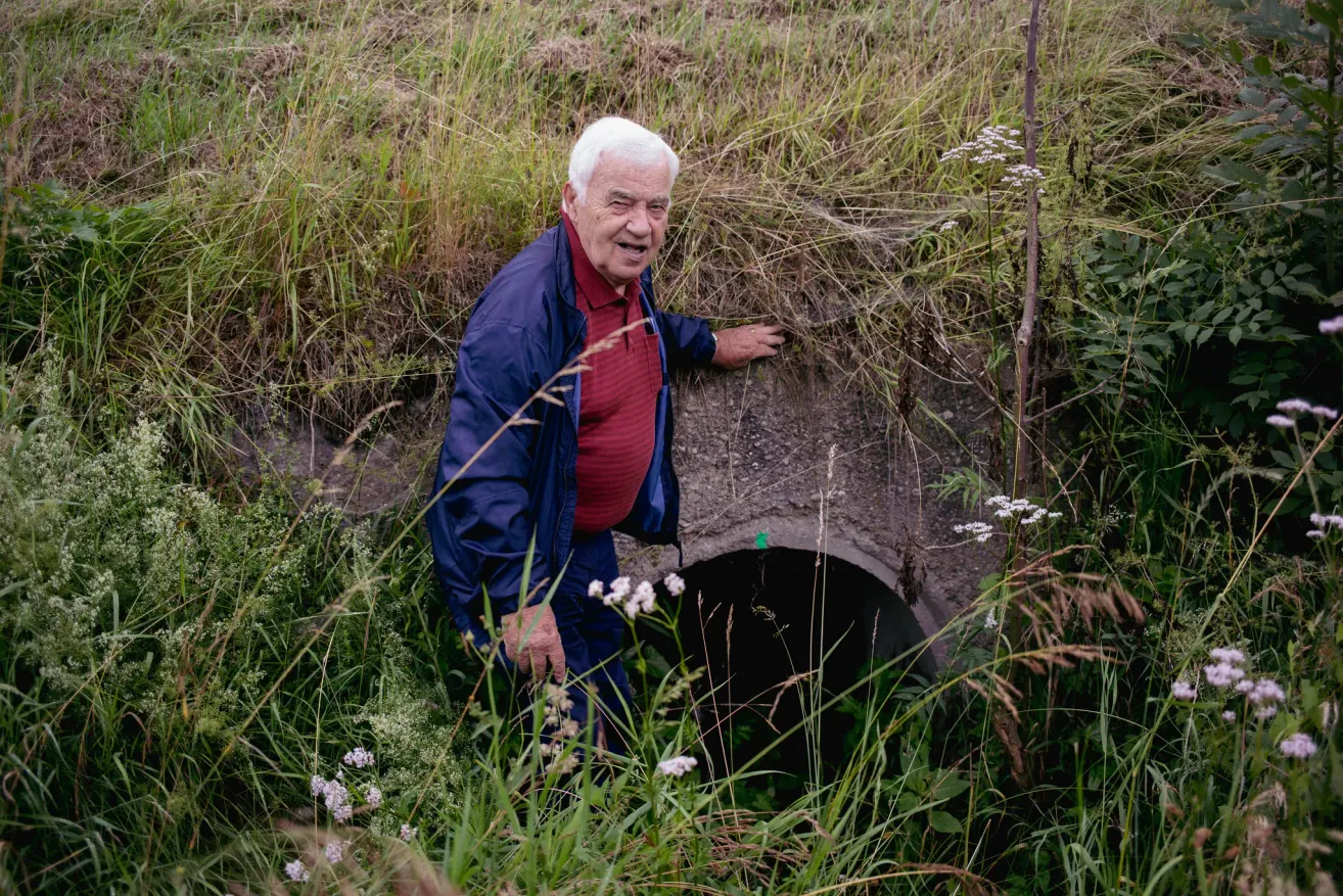 Ein älterer Mann in blauer Jacke und rotem Hemd steht neben einem runden Loch in einem Grasfeld.