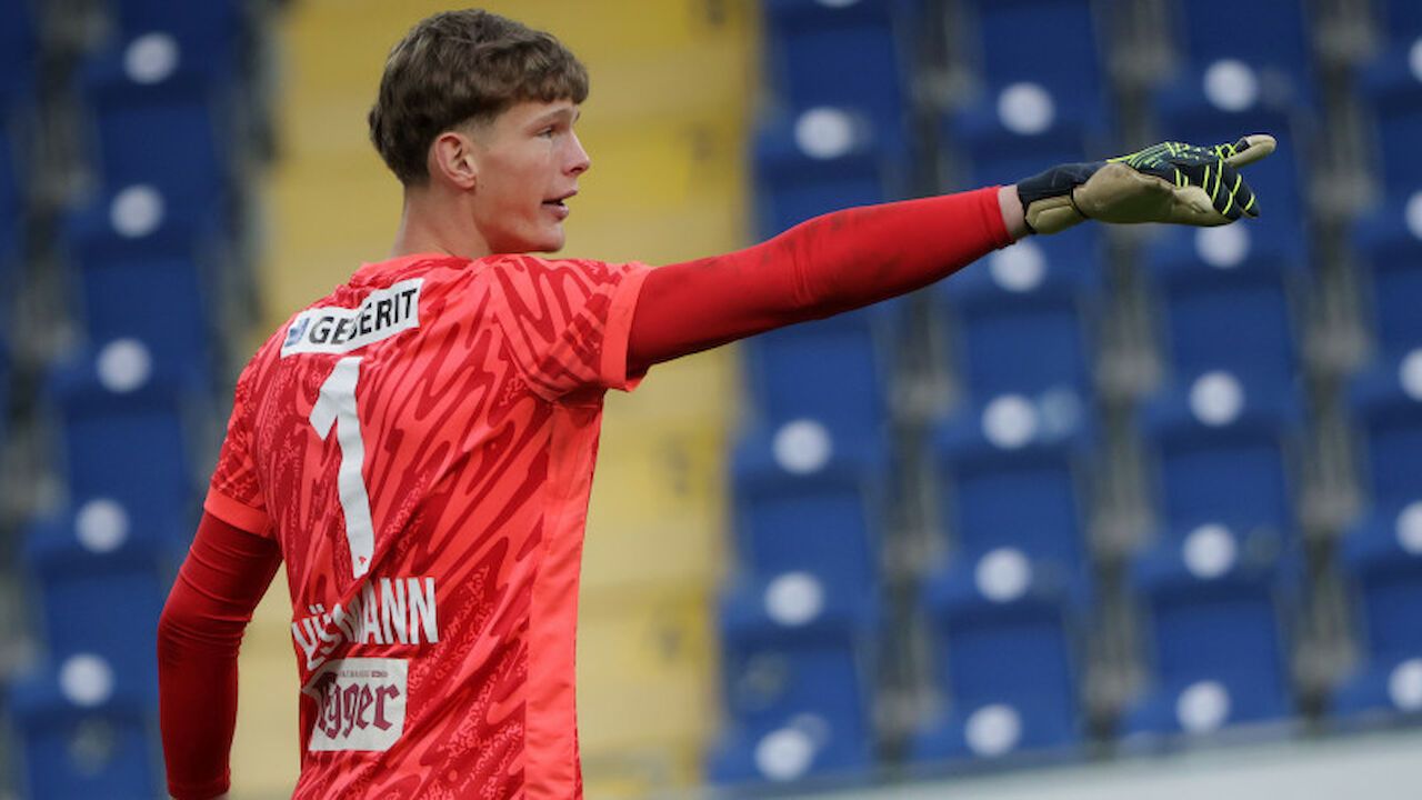 Ein Fußballspieler in einem roten Trikot mit 'GEERI'T' und der Nummer '1', steht in einem Stadion mit leeren blauen Sitzen.
