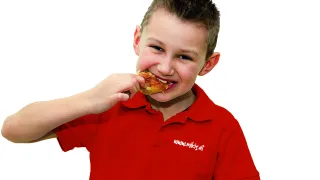 Ein junger Junge in einem roten Poloshirt mit Logo isst mit den Händen Huhn. Er lächelt und sieht zufrieden aus.