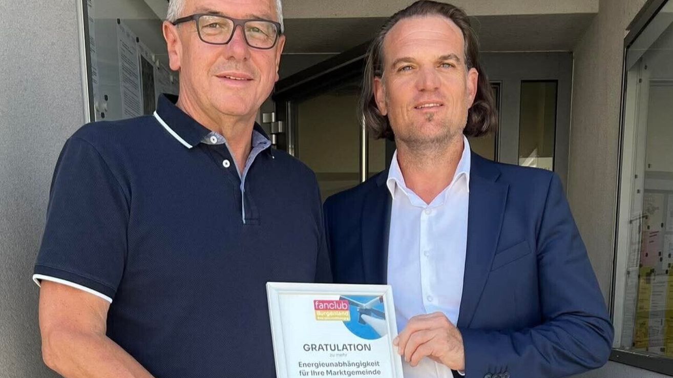 Zwei Männer stehen nebeneinander und lächeln, einer hält ein Zertifikat für Energieunabhängigkeit. Der Mann auf der linken Seite trägt eine Brille und ein marineblaues Poloshirt, während der Mann auf der rechten Seite einen marineblauen Blazer trägt.