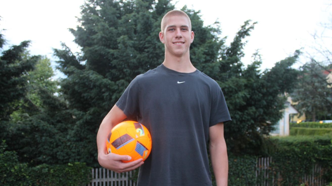 Ein Mann in einem grauen Nike-Shirt hält einen orangefarbenen Fußball. Er steht vor einem grünen Baum und lächelt.