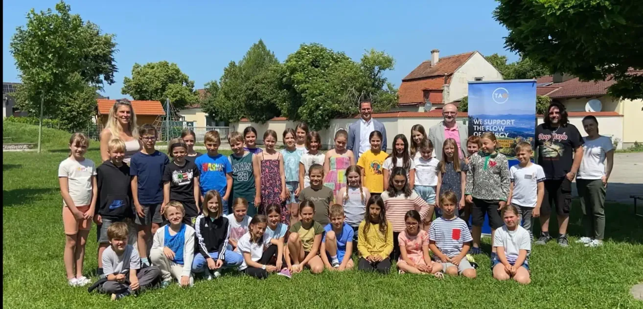 Eine Gruppe von Kindern und zwei Erwachsenen posiert für ein Foto in einem Park. Sie sitzen und stehen auf dem Rasen mit Gebäuden im Hintergrund.