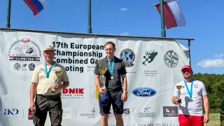 Drei Männer stehen auf einem Podium mit Medaillen um den Hals. Einer hält eine Trophäe. Hinter ihnen steht ein Banner für die 17. European Championship Combined Game Shooting. Flaggen hängen an Stangen im Hintergrund.