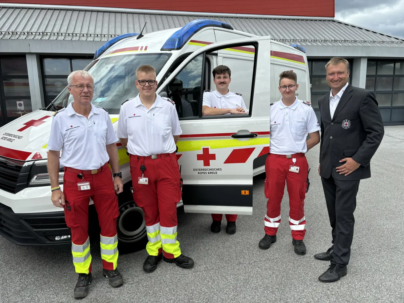 Vier Männer in roten und weißen Uniformen stehen vor einem Krankenwagen. Der Mann in der Mitte ist im Fahrzeug und lächelt. Der Krankenwagen hat ein rotes Kreuz und den Text 'Deutsches Rotes Kreuz' an seiner Seite.