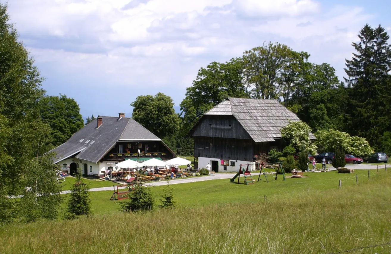 Zwei rustikale Häuser stehen auf einem Rasen mit Bäumen und Büschen. Ein Spielplatz befindet sich in der Nähe des größeren Hauses, unter dem Menschen unter Schirmen sitzen. Eine Person geht am kleineren Haus vorbei.