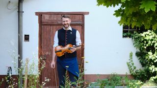 Ein Mann in einer blauen Schürze und Brille lächelt, hält eine Geige und steht vor einer Holztür mit einer weißen Wand dahinter.