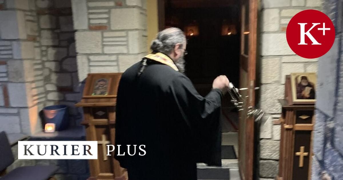 Ein Priester in einem schwarzen Gewand steht vor einer Kirchentür und hält einen Schlüssel. Der Raum ist schwach beleuchtet.