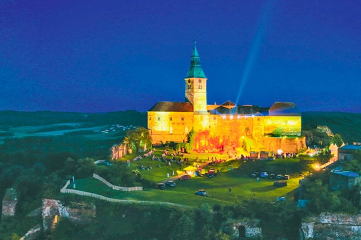 Eine Luftaufnahme einer mittelalterlichen Burg bei Nacht, beleuchtet mit gelben Lichtern. Es gibt Menschen und Autos auf dem Rasenfeld. Bäume und Berge sind im Hintergrund.