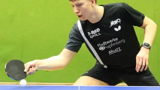 Ein junger Mann spielt Tischtennis auf einem blauen Tisch mit grünem Hintergrund, trägt ein schwarzes Shirt mit weißen Logos und eine Uhr.