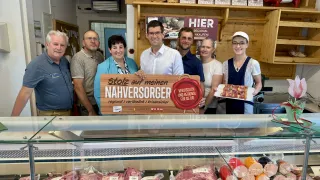 Eine Gruppe von Menschen steht hinter einer Fleischtheke in einem Geschäft. Sie lächeln und posieren für ein Foto. Die Theke hat verschiedene Fleischsorten. Das Schild sagt 'Stolza auf meinen nahversdorfer regional verlasslich krinser.' Die Leute tragen Freizeitkleidung.