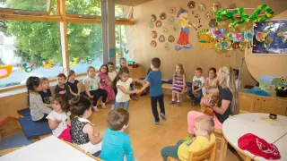 Eine Gruppe von Kindern sitzt im Kreis um eine Frau, die eine Gitarre spielt. Die Kinder beschäftigen sich mit verschiedenen Aktivitäten, einschließlich des Zuhörens der Frau. Der Raum hat Holzböden und Wände mit verschiedenen Gemälden.