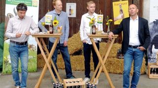 Vier Männer stehen hinter Tischen, die mit Flaschen von Frastanzer-Bier gefüllt sind. Ein Mann hält eine Flasche und ein anderer Mann spricht. Sie sind von Heuballen umgeben.