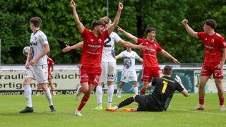 Fußballspieler in Rot feiern ein Tor, ein Spieler in Weiß fällt auf den Boden, umgeben von Spielern in weißen und roten Trikots.
