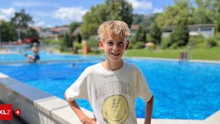 Ein junger Junge steht neben einem Schwimmbecken, lächelt und posiert für ein Foto. Er trägt ein weißes T-Shirt mit einem gelben Smiley-Gesicht. Hinter ihm befindet sich ein großes, blaues Becken, und in der Ferne sind Bäume und Gebäude zu sehen.
