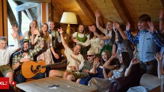 Eine Gruppe von Menschen in traditioneller Kleidung ist in einem Raum, einige sitzen auf einer Couch und einige stehen. Eine Person hält eine Gitarre und spielt sie. Sie heben alle ihre Hände und scheinen sich zu amüsieren.