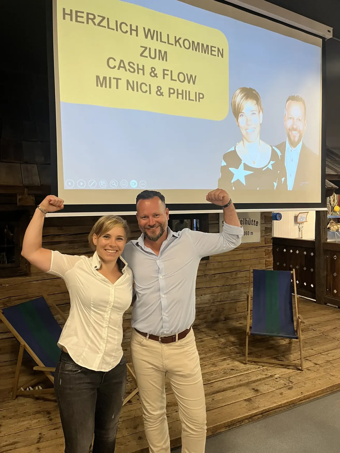 Ein Mann und eine Frau stehen lächelnd vor einer Projektionsleinwand mit den Worten 'Cash & Flow mit Nici & Philip'. Sie heben ihre Arme und scheinen sich in einem professionellen Umfeld zu befinden.