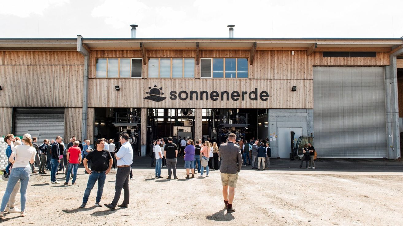 Eine Gruppe von Menschen steht vor einem modernen Gebäude namens 'Sonnenerde', mit einem Sonnenlogo und einem Gabelstapler in der Nähe.