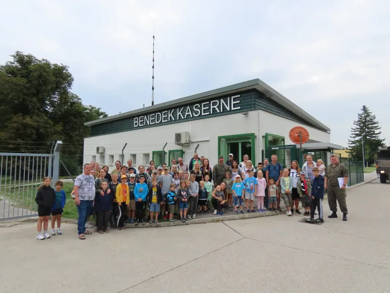 Eine große Gruppe von Menschen steht vor dem Gebäude der Benedek Kaserne, mit Bäumen und einem klaren Himmel im Hintergrund.