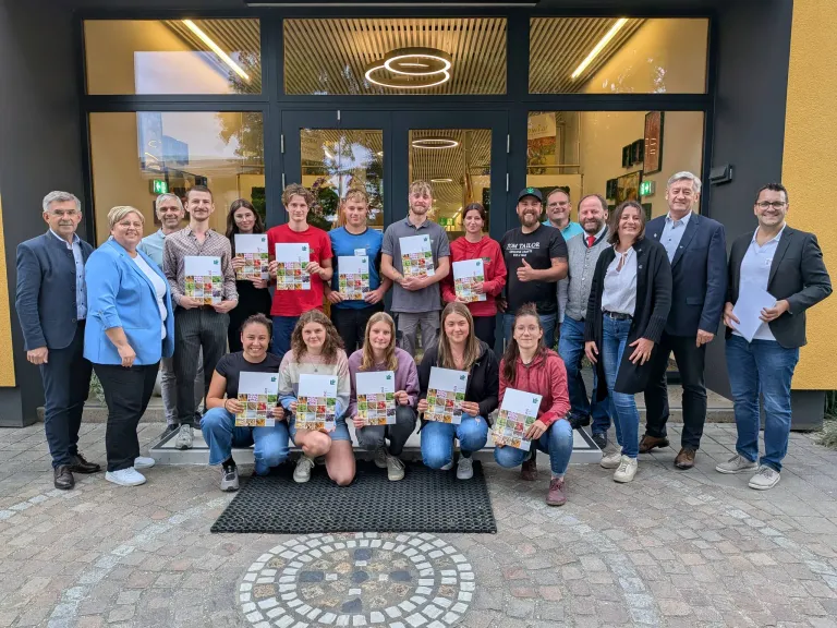 Eine Gruppe von Menschen in Freizeitkleidung posiert für ein Foto vor einem modernen Gebäude und hält Urkunden in den Händen. Sie lächeln und scheinen eine Errungenschaft zu feiern.