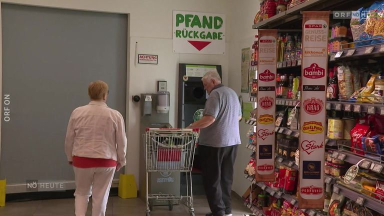 Zwei Personen befinden sich in einem Geschäft. Ein älterer Mann hält einen Einkaufswagen. Eine Frau steht hinter ihm. Flaschen und Snacks sind in den Regalen.