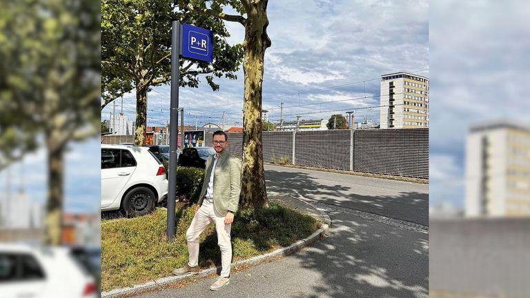 Ein Mann steht an einer Parkzeichen an einer Straßenecke, trägt eine Brille und einen hellgrünen Anzug. Hinter ihm steht ein Baum, und im Hintergrund sind mehrere Gebäude zu sehen.