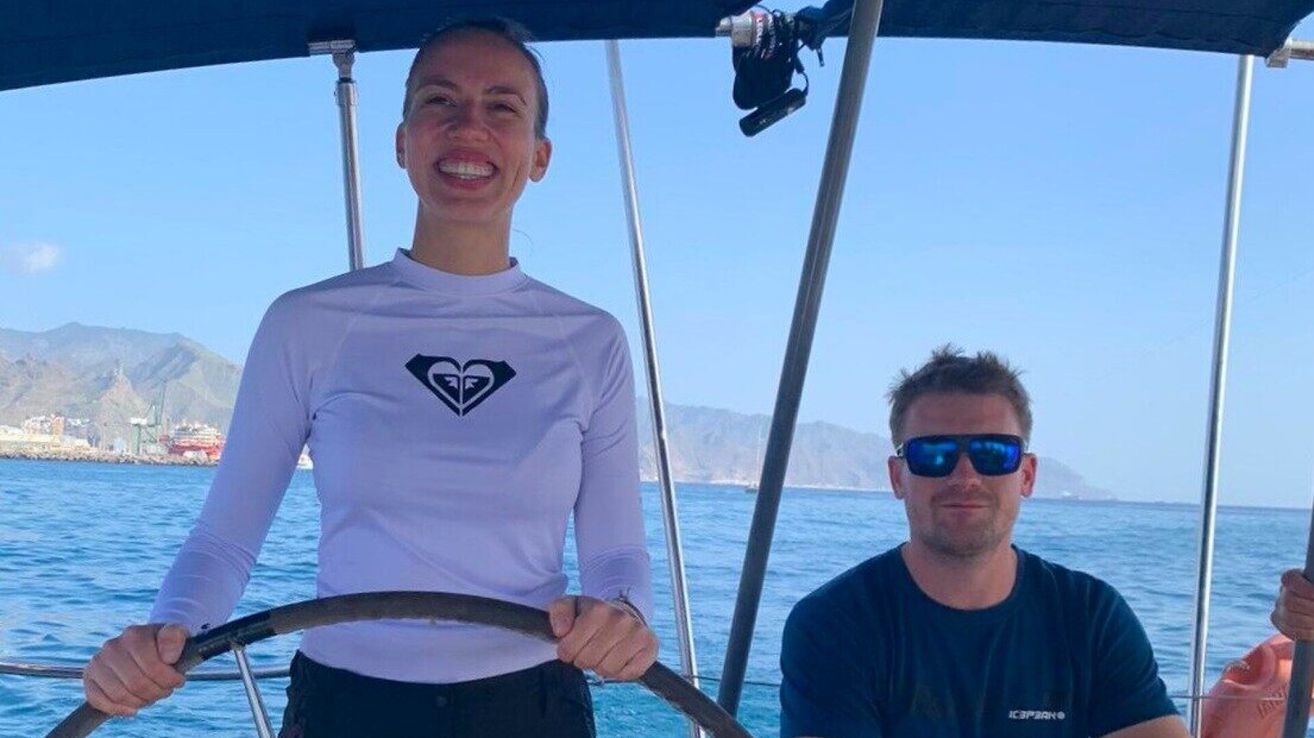 Eine Frau und ein Mann auf einem Boot, wobei die Frau das Steuerrad hält und lächelt. Der Mann trägt eine Sonnenbrille. Sie befinden sich an einem sonnigen Tag mit Bergen im Hintergrund.