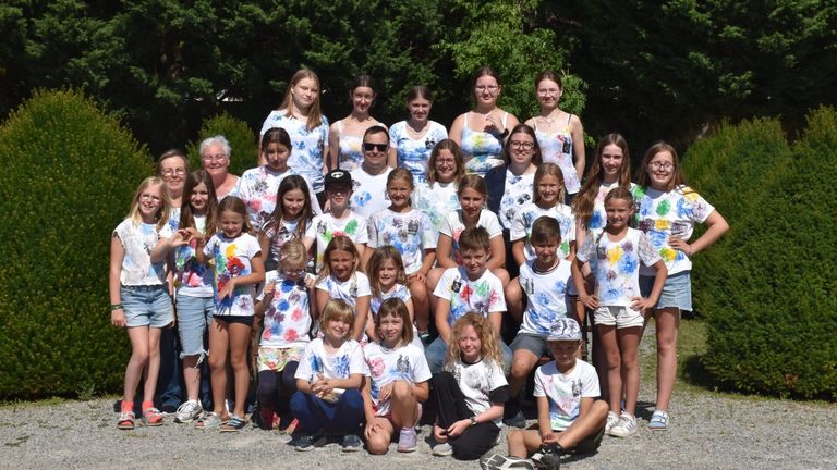 Eine Gruppe junger Kinder und Teenager, alle in Tie-Dye-Shirts gekleidet, posiert für ein Foto im Freien. Bäume sind im Hintergrund zu sehen.