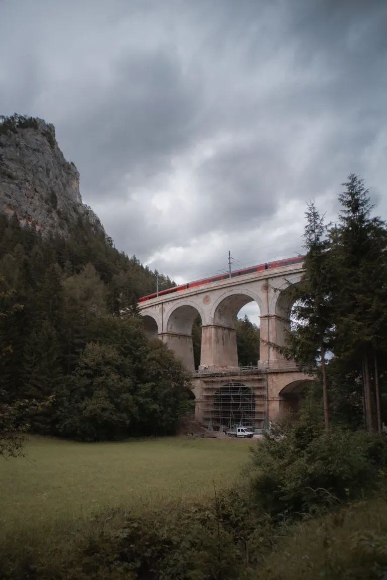 Ein roter Zug überquert eine große Steinbrücke in einem bergigen Gebiet mit Bäumen und einem bewölkten Himmel.
