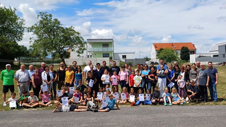 Eine Gruppe von Menschen, Erwachsene und Kinder, stehen und sitzen draußen, halten Papiere in der Hand, mit Gebäuden im Hintergrund unter einem teilweise bewölkten Himmel.