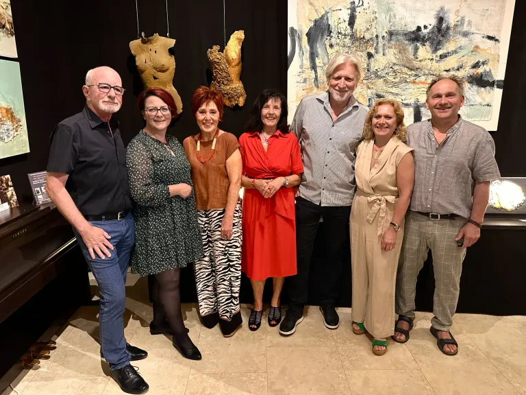 Eine Gruppe von Menschen steht vor einer Kunstgalerie. Sie lächeln und scheinen für ein Foto zu posieren. Hinter ihnen befinden sich abstrakte Gemälde und Skulpturen.