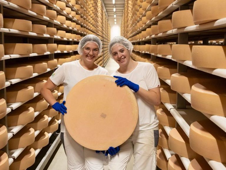 Zwei Frauen in weißen Uniformen und Haarnetzen halten ein großes Käserad in einem Lagerraum, der mit gestapelten Käseblöcken gefüllt ist.