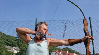 Ein Mann steht auf einem Feld und bereitet sich darauf vor, einen Pfeil mit einem Bogen abzuschießen. Er trägt ein ärmelloses Hemd und Handschuhe. Hinter ihm befindet sich ein Zaun mit Häusern und Bäumen.