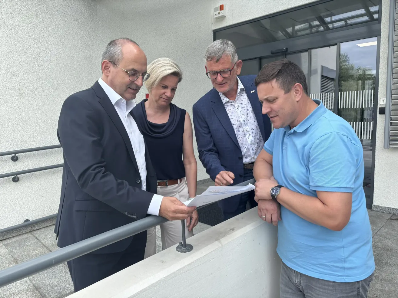 Vier Personen, drei Männer und eine Frau, betrachten ein Stück Papier auf einer Brüstung. Der Mann auf der linken Seite hält das Papier, während der Mann auf der rechten Seite die Hände verschränkt hat.