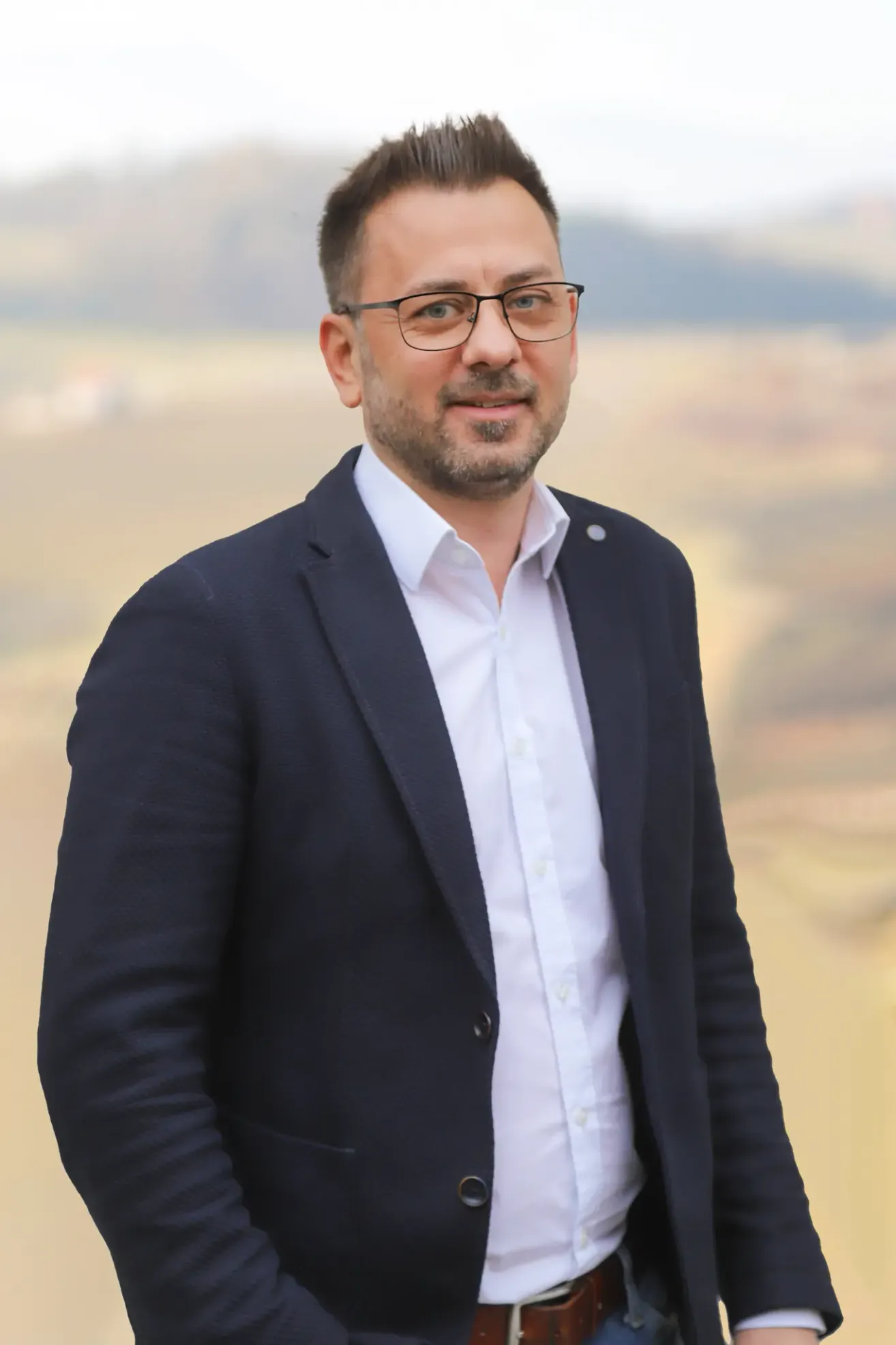 Ein Mann mit Bart und Brille steht lächelnd und trägt ein weißes Hemd und einen marineblauen Blazer mit einer Anstecknadel auf der linken Seite.