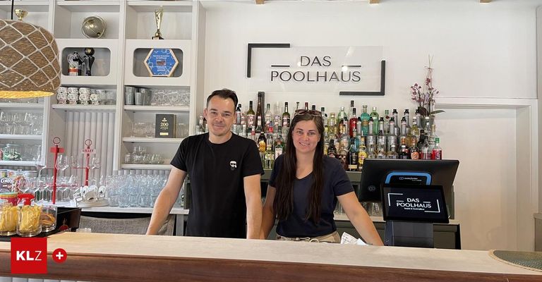 Zwei Menschen stehen hinter einer Bar mit einer großen Auswahl an Alkohol, einer lächelt und der andere hat eine Sonnenbrille auf dem Kopf. Über ihnen steht ein Schild mit der Aufschrift 'DAS POOLHAUS'.