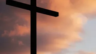 Eine Silhouette eines Kreuzes steht vor einem Hintergrund eines Himmels voller Wolken. Das Kreuz ist dunkel und kontrastiert mit den orangen und rosa Tönen des Himmels.