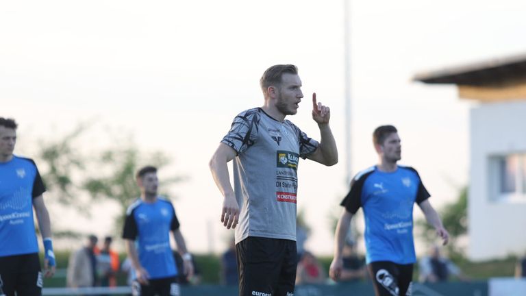 Ein Fußballspieler in einem grauen Trikot mit schwarzen Shorts gibt auf einem Rasenfeld eine Geste von sich, während zwei andere Spieler in blauen Trikots im Hintergrund stehen.