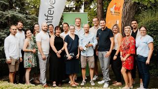 Eine Gruppe von Menschen steht in einem Park zusammen, lächelt und posiert für ein Foto. Sie sind lässig gekleidet, und eine Person hält eine Flasche Wein. Hinter ihnen befinden sich Banner und ein Surfbrett mit den Buchstaben 'JW' darauf. Im Hintergrund sind Bäume zu sehen.