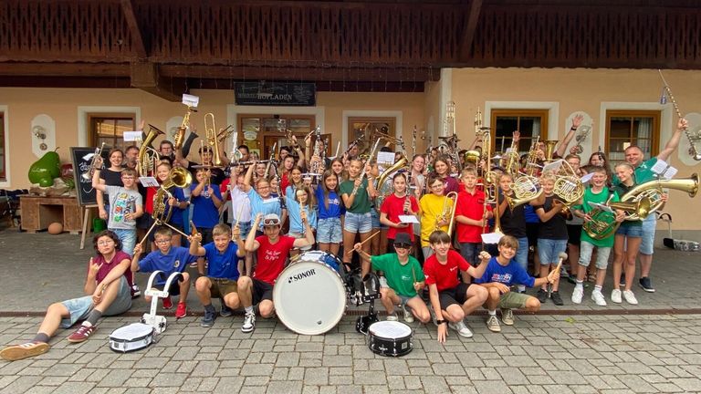 Eine Gruppe junger Musiker posiert mit verschiedenen Instrumenten vor einem Gebäude. Sie sind in Freizeitkleidung gekleidet und einige tragen Brillen. Hinter ihnen steht ein Schild mit der Aufschrift 'MÖLLZU HOTLADEN'.